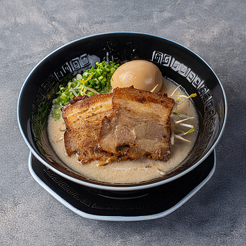 검은 그릇에 담긴 일본식 돈코츠 라멘으로, 크리미한 국물 위에 구운 차슈 고기 두 장, 반숙 달걀, 숙주와 다진 파가 올려진 모습입니다.