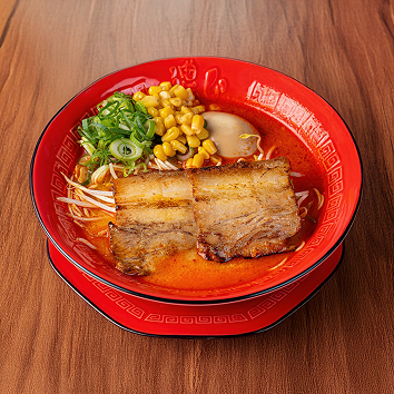 붉은 그릇에 담긴 일본식 라멘으로, 매콤해 보이는 붉은 국물 위에 차슈 고기 두 장, 옥수수, 숙주, 다진 파가 올려져 나무 테이블 위에 놓인 모습입니다.